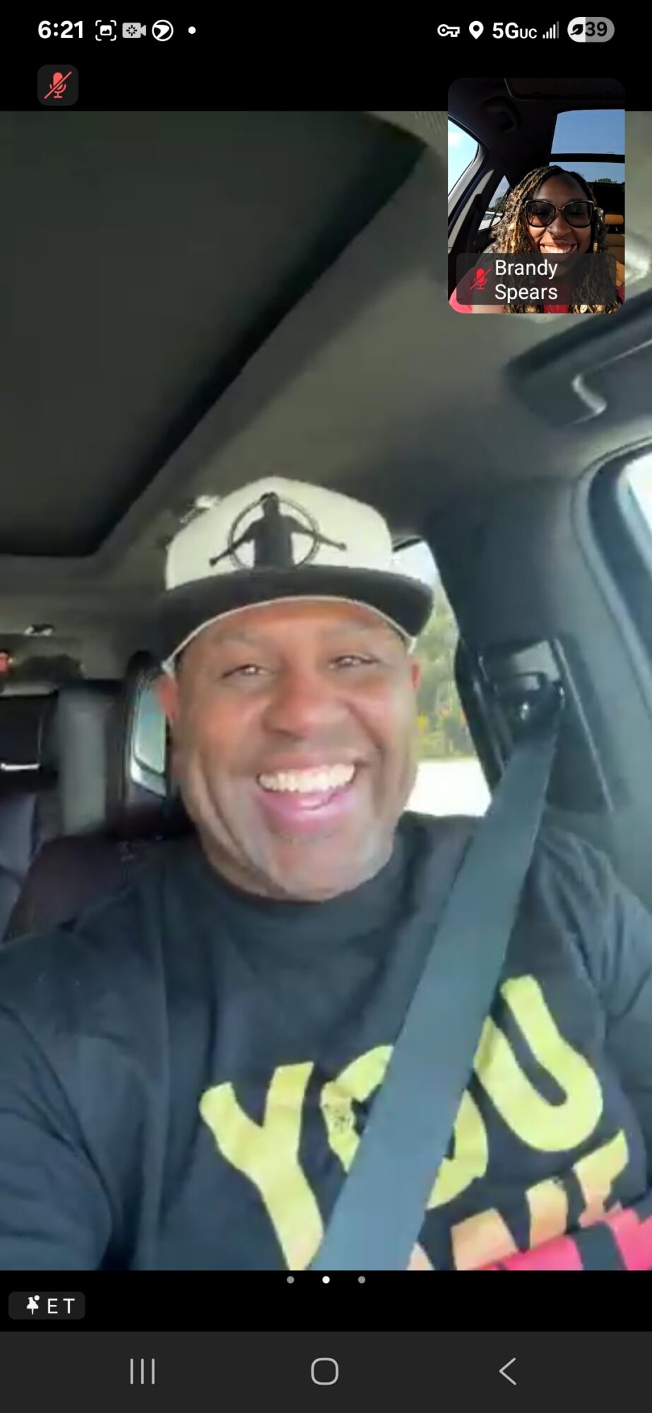 Smiling man wearing a cap and seatbelt inside a vehicle, with a shadow of a drone on the ceiling, during a video call.