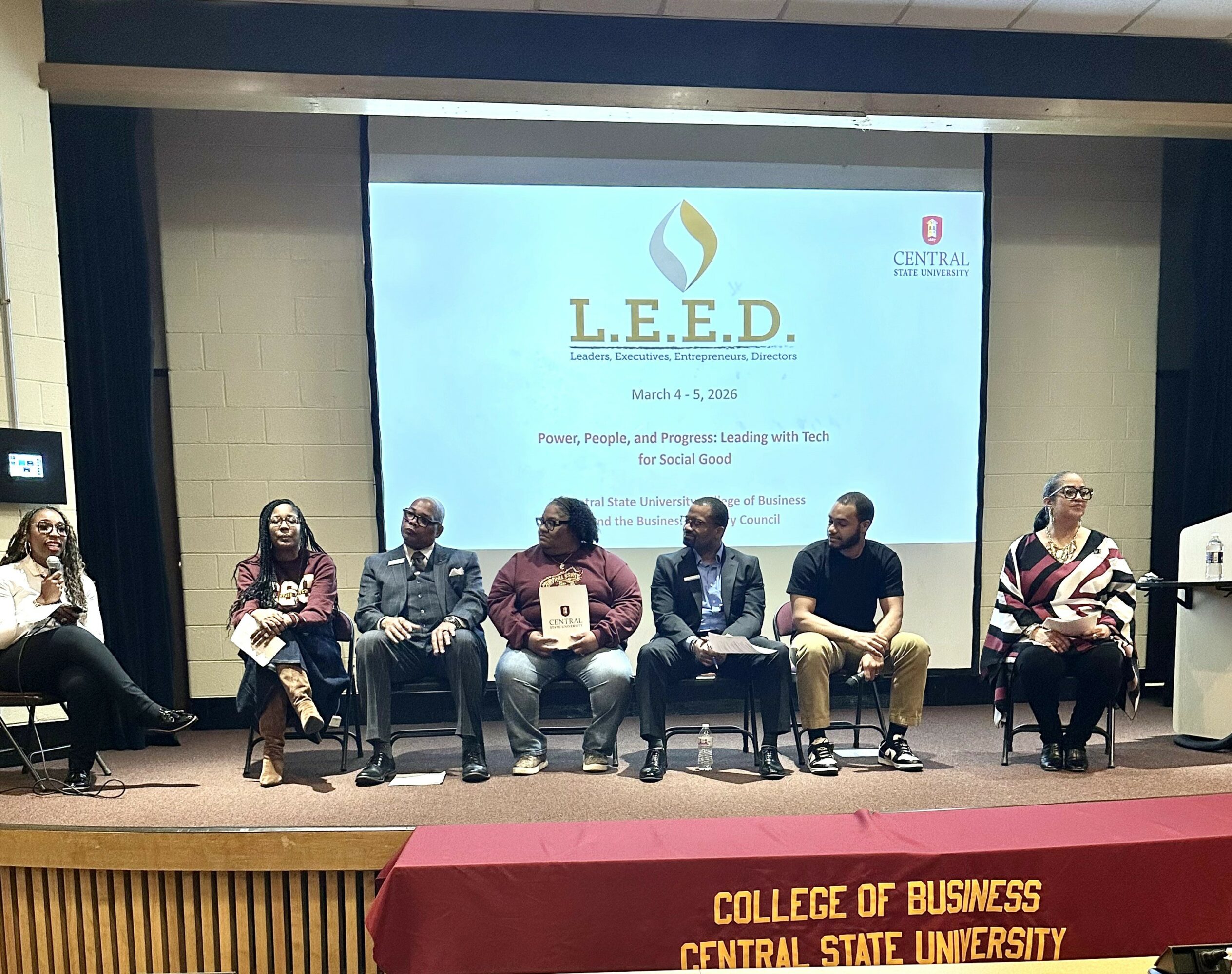 Six people seated on stage in front of a large screen displaying a presentation titled 'L.E.E.D.' at a college event.