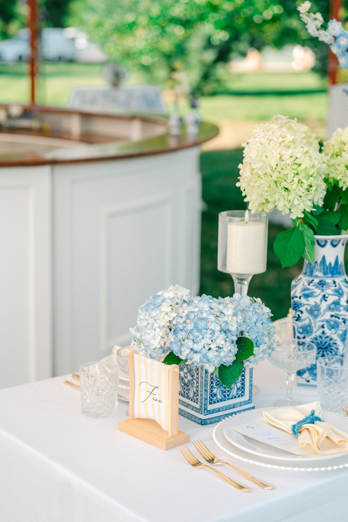 Table decorated with flowers, candles, and tableware outdoors with greenery in background.