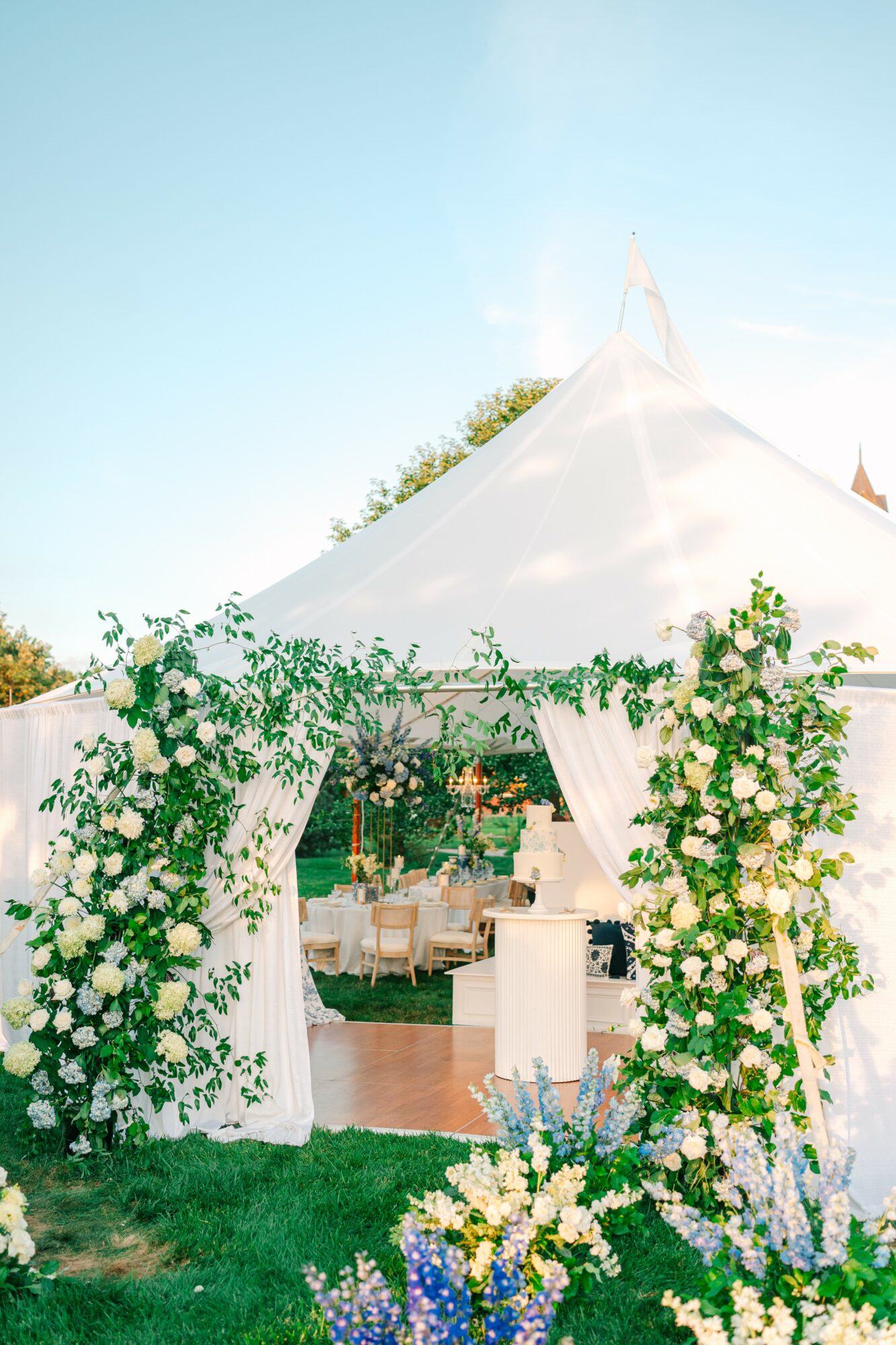 White tent with floral arch entrance, outdoor setting with tables and chairs inside, blue sky above.