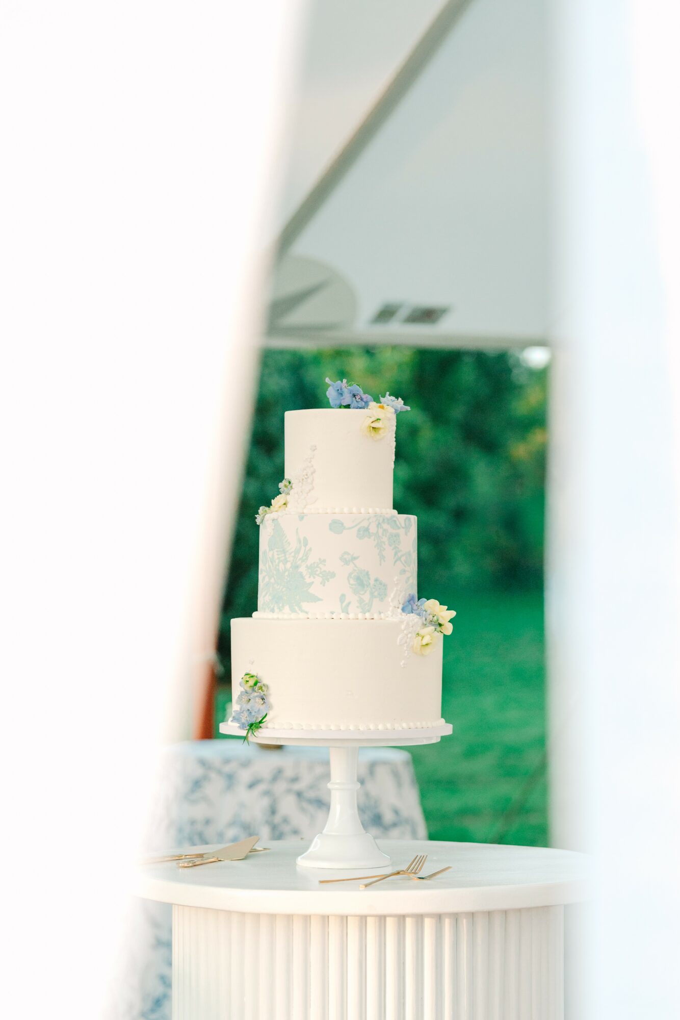 Three-tiered white wedding cake with floral decorations on a cake stand outdoors, viewed through a narrow opening.