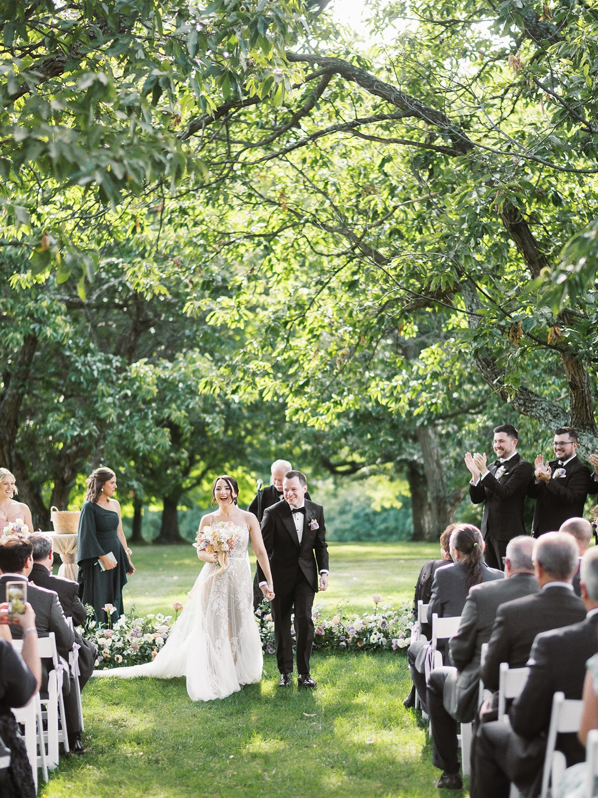 A wedding ceremony outdoors with a bride and groom walking on grass, surrounded by seated guests and lush green trees.