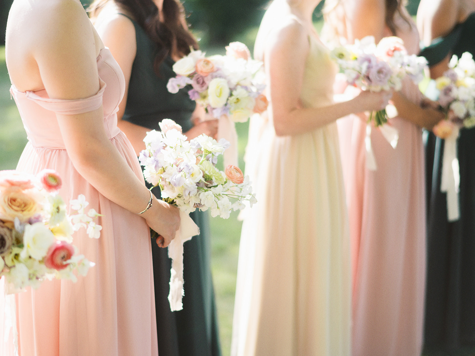 Group of women in pastel and dark dresses holding bouquets at an outdoor event.