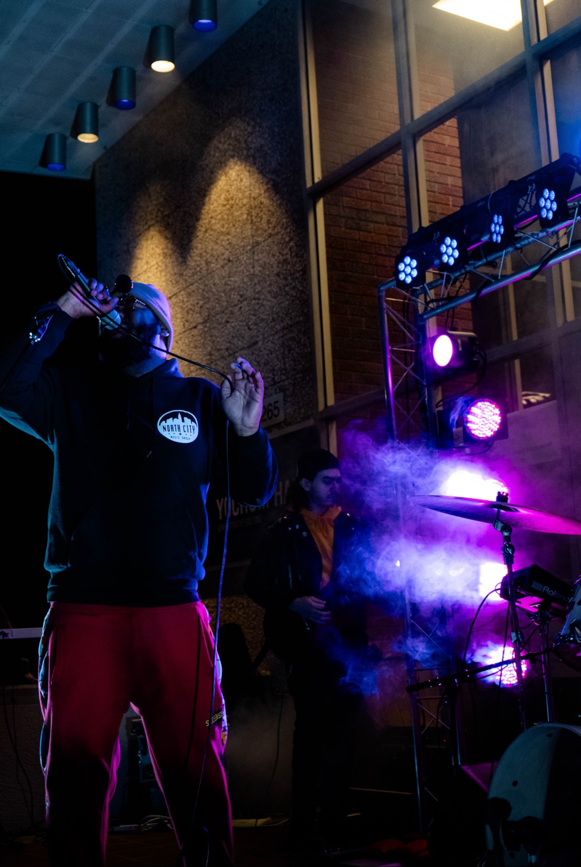 Person singing into a microphone on stage with purple lighting and smoke effects, in a dimly lit indoor venue.