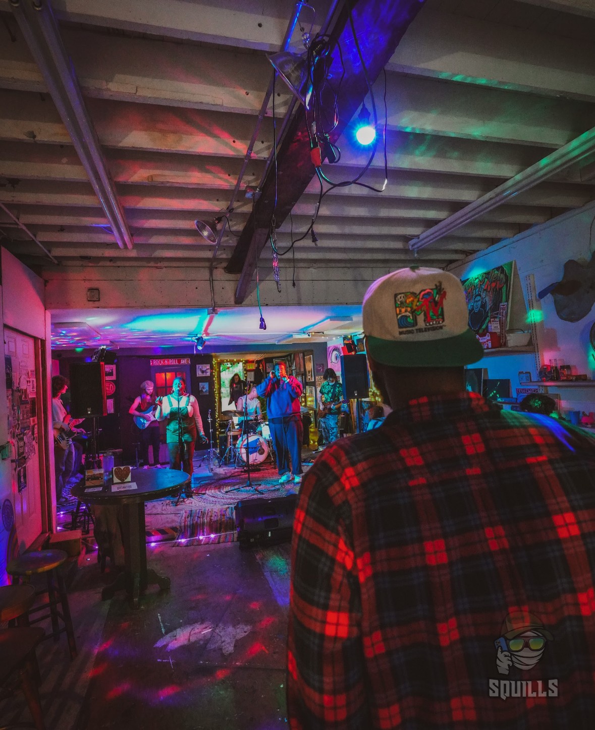 Person wearing a plaid shirt and cap watching a live music performance in a colorful, indoor venue.