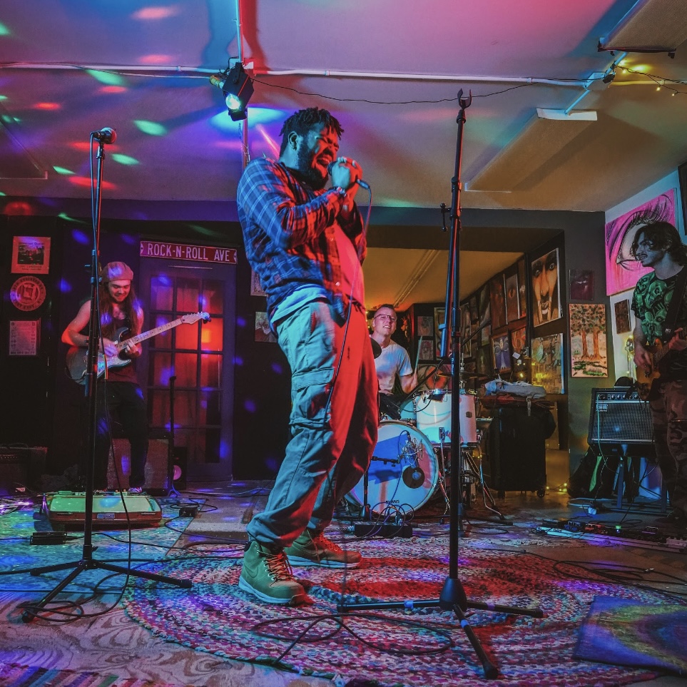 A band performs on stage with colorful lighting, featuring a singer, guitarist, bassist, and drummer in a cozy venue.