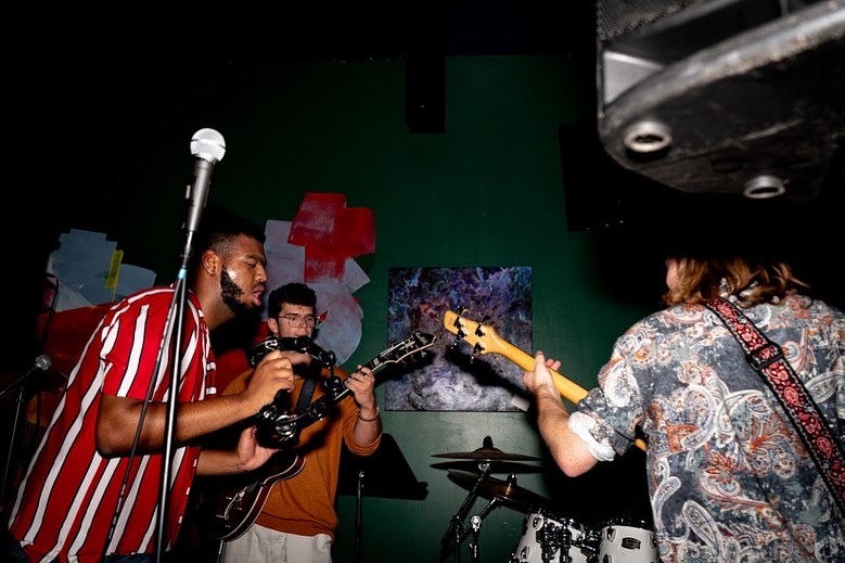 Three musicians perform on stage, one playing guitar, another with a camera, and a third with a microphone, in a dimly lit setting.