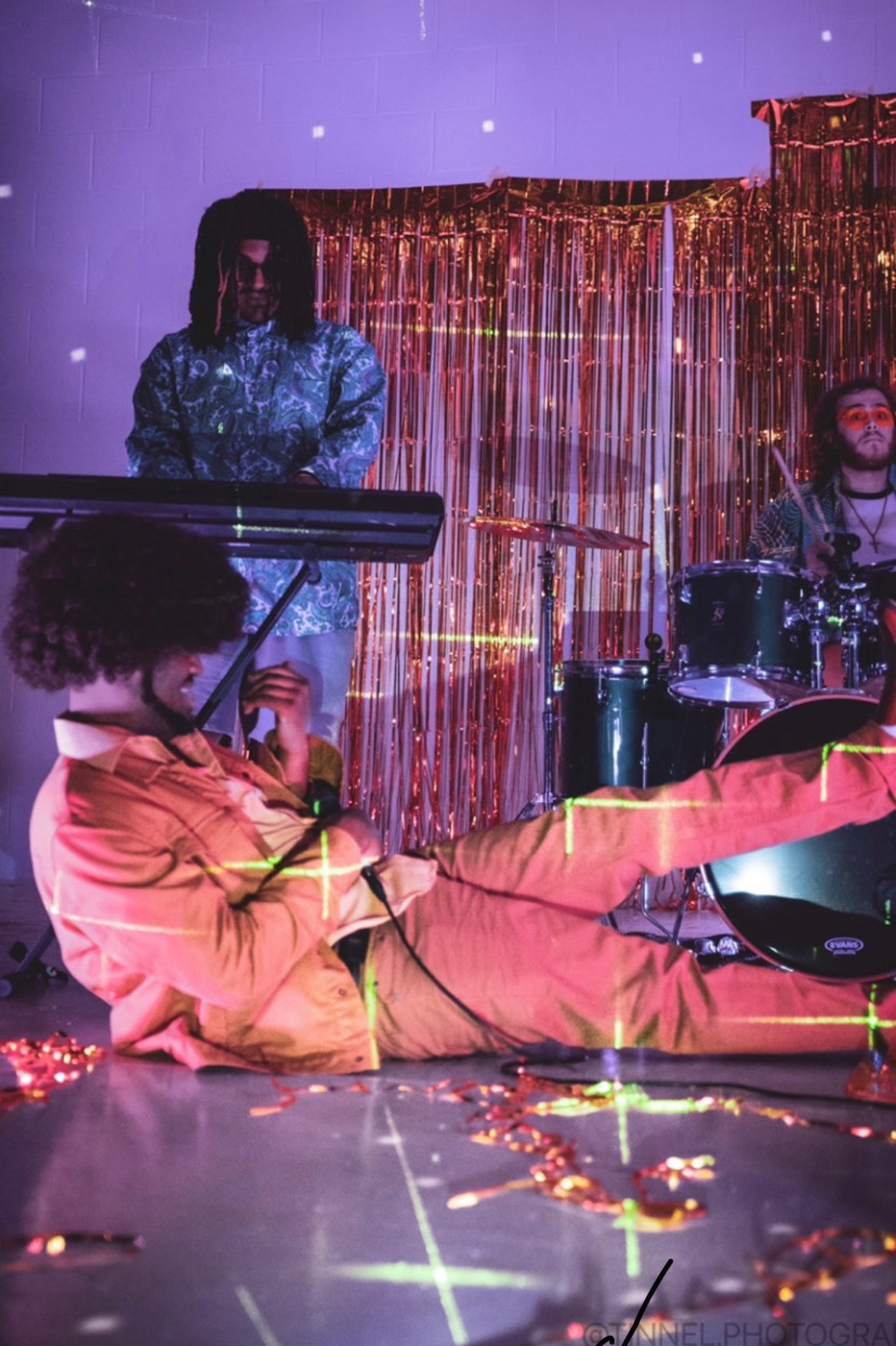 Person lying on the floor playing a violin, with a drummer and keyboardist performing in the background, stage decorated with shiny curtains.