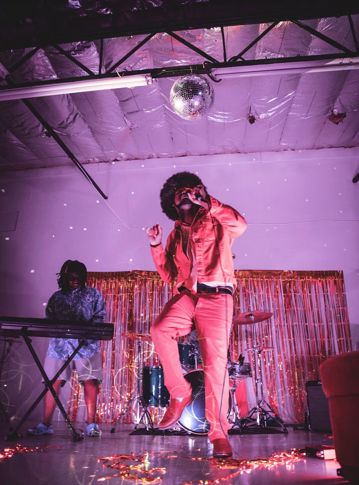 Person dancing on stage with a microphone, a child playing keyboard, and musical instruments in the background, under purple lighting.