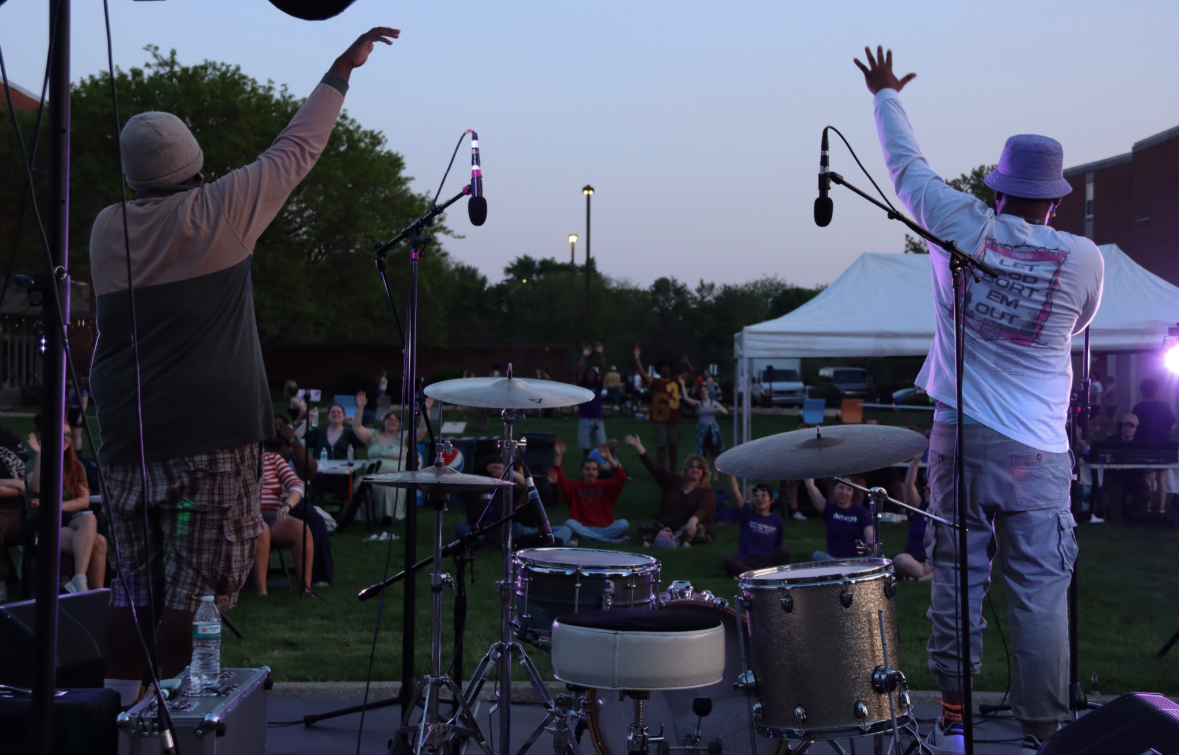 Two performers on stage with drums and microphones, raising their hands, audience seated outdoors at dusk.