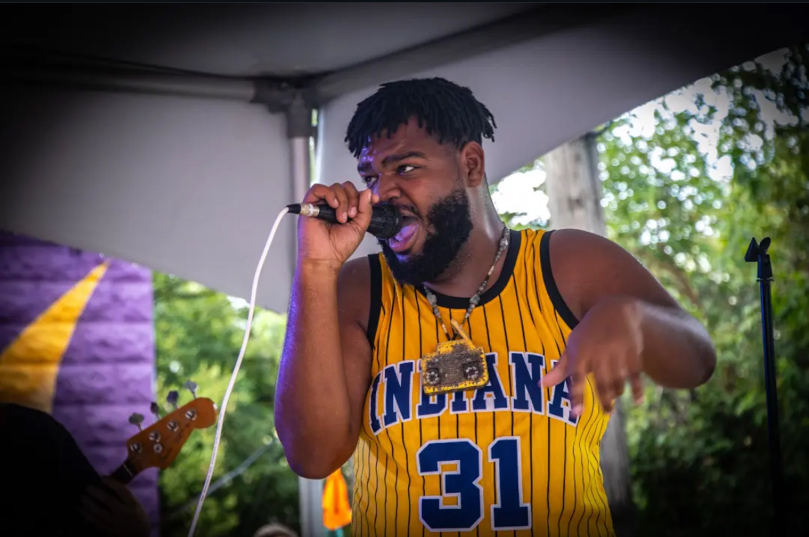 Person singing into a microphone outdoors, wearing a yellow basketball jersey with the number 31, with trees in the background.