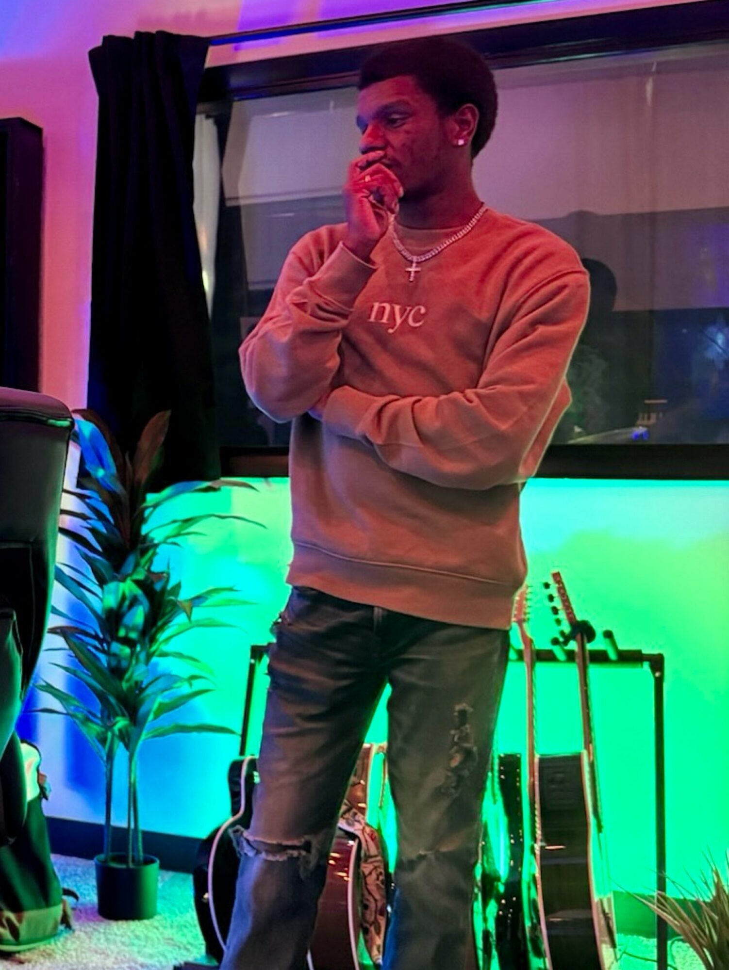 Person standing indoors with hand near face, wearing a beige sweatshirt and ripped jeans, illuminated by colorful lights.