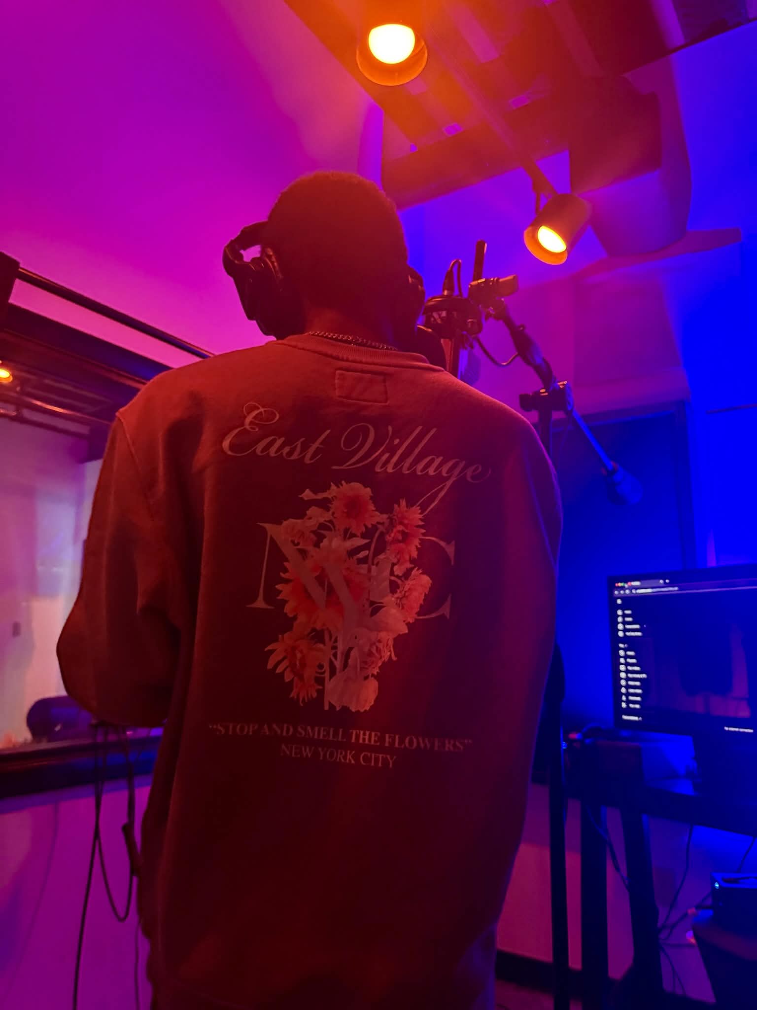 Person with headphones in a colorful, dimly lit room with equipment and a computer screen, facing away from the camera.