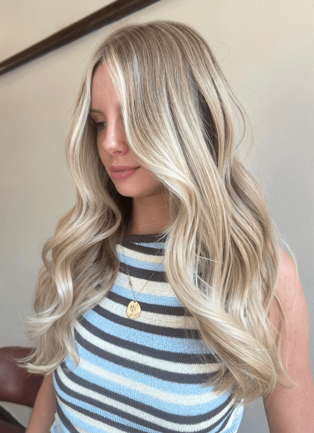 Woman with long, wavy blonde hair wearing a striped sleeveless top and a gold necklace, looking down.