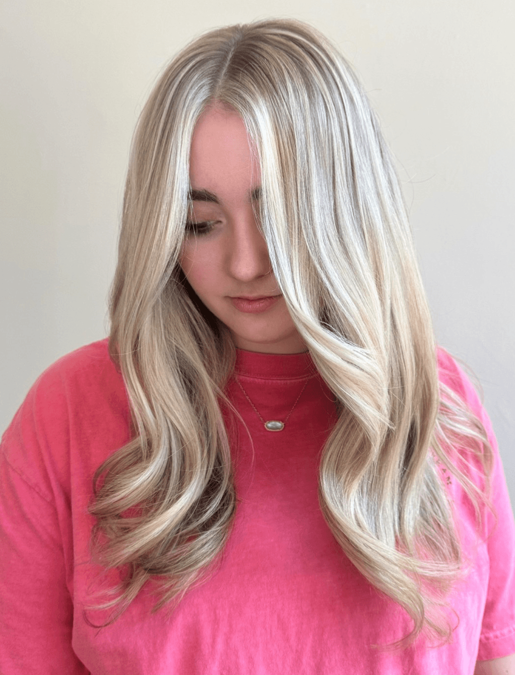 Young woman with long, wavy blonde hair wearing a pink top, looking downward, neutral background.