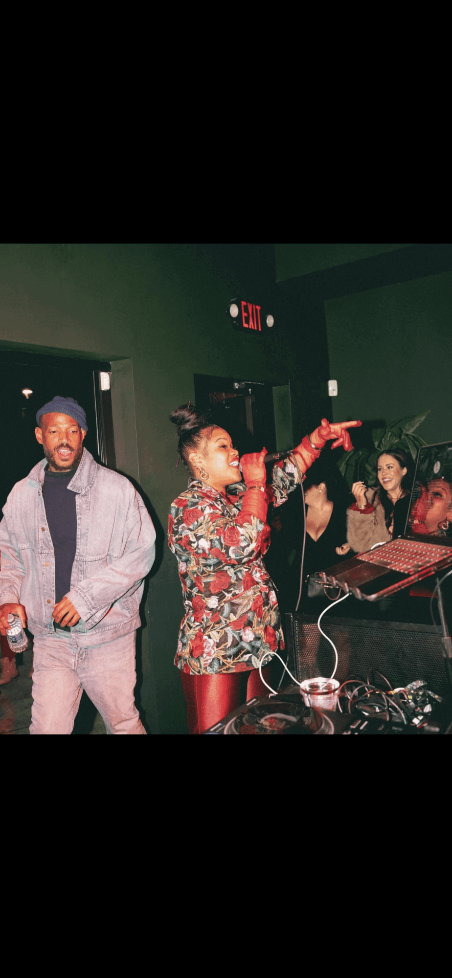 People performing music and singing in a dimly lit room with equipment and a green wall.