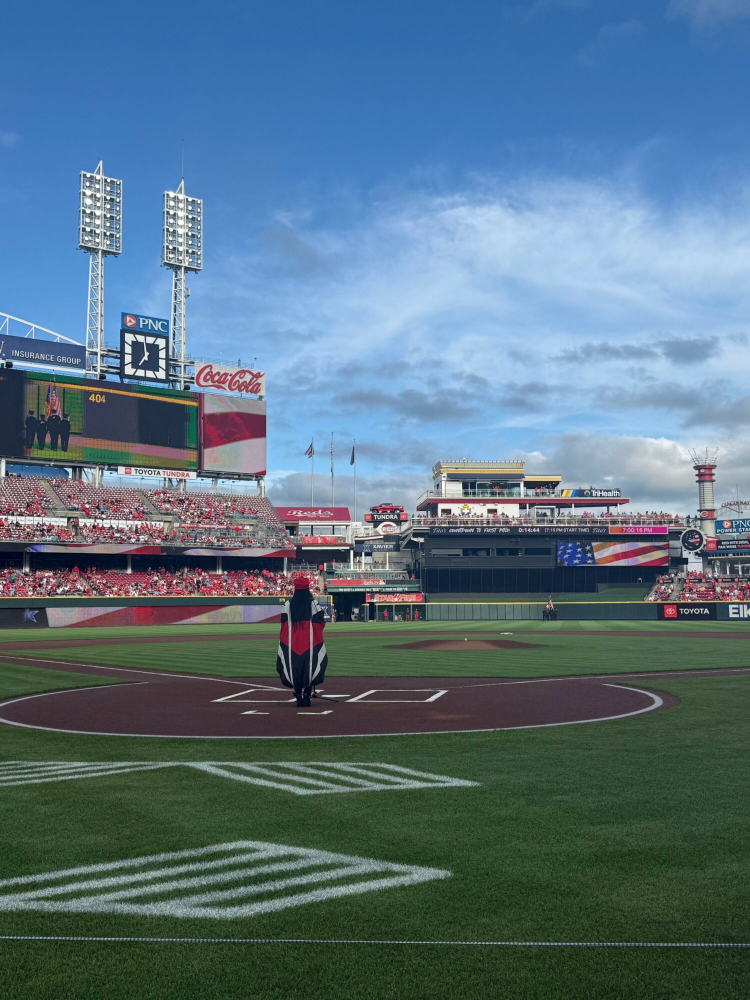 Baseball field with a person standing near home plate, stadium, and sky in the background.