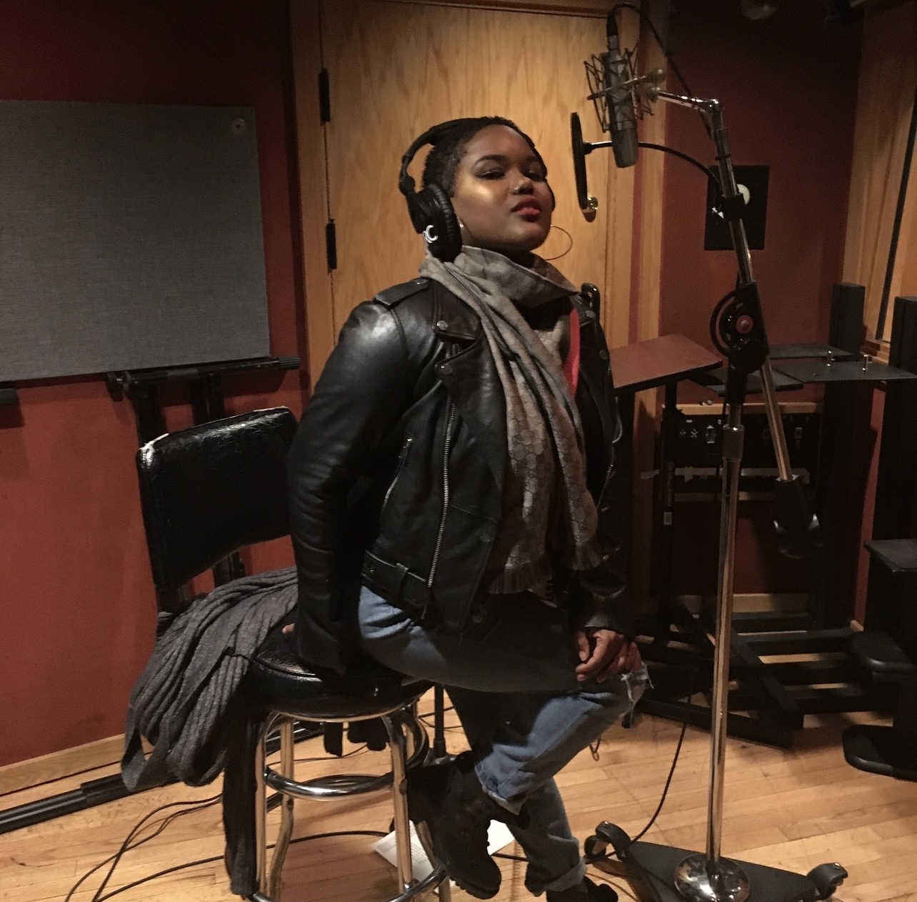 Woman in a recording studio wearing headphones, leather jacket, and scarf, sitting on a stool with a microphone.
