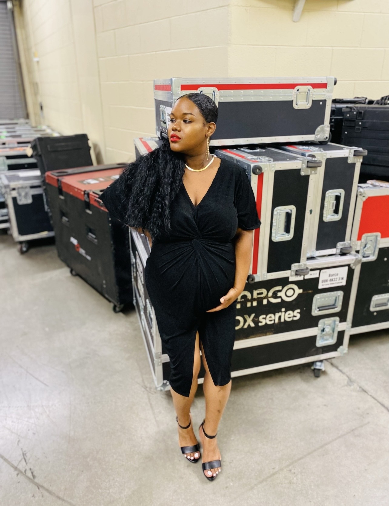 Woman in black dress with long curly hair standing in backstage area with equipment cases behind her.