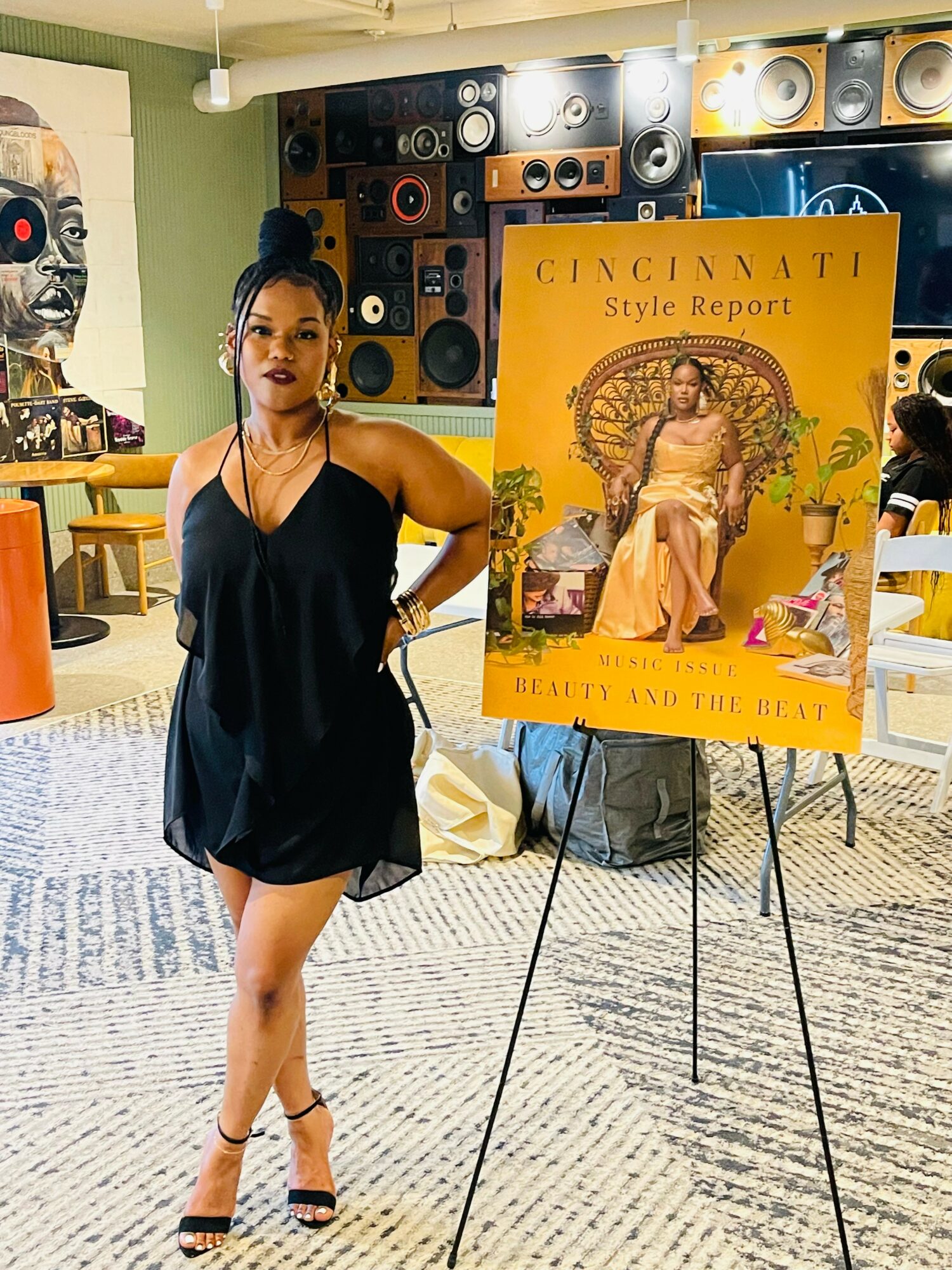 Woman in black dress standing next to a yellow Cincinnati Style Report poster at an indoor event.