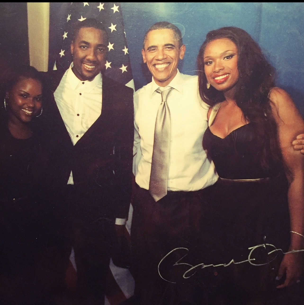 Four people smiling, standing in front of an American flag, dressed in formal attire, with a blue background.