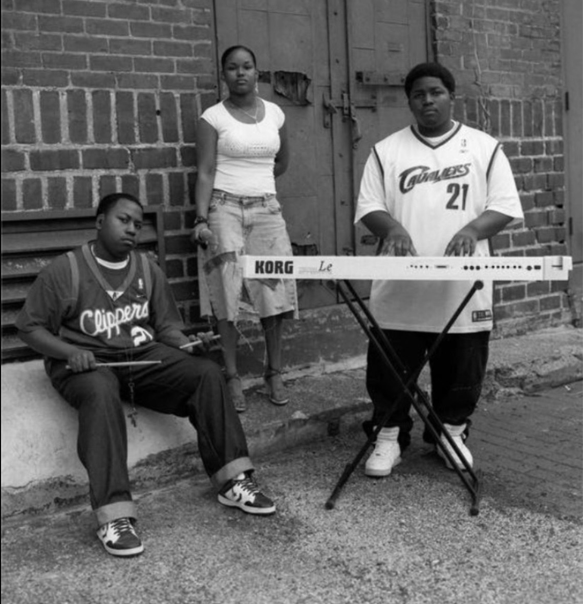 Three young people pose outdoors with a keyboard and a brick wall background.