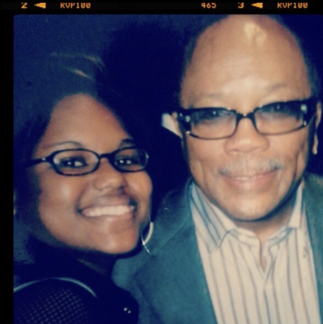 Two people smiling, wearing glasses, close together, dark background, one with striped shirt, the other with dark clothing.