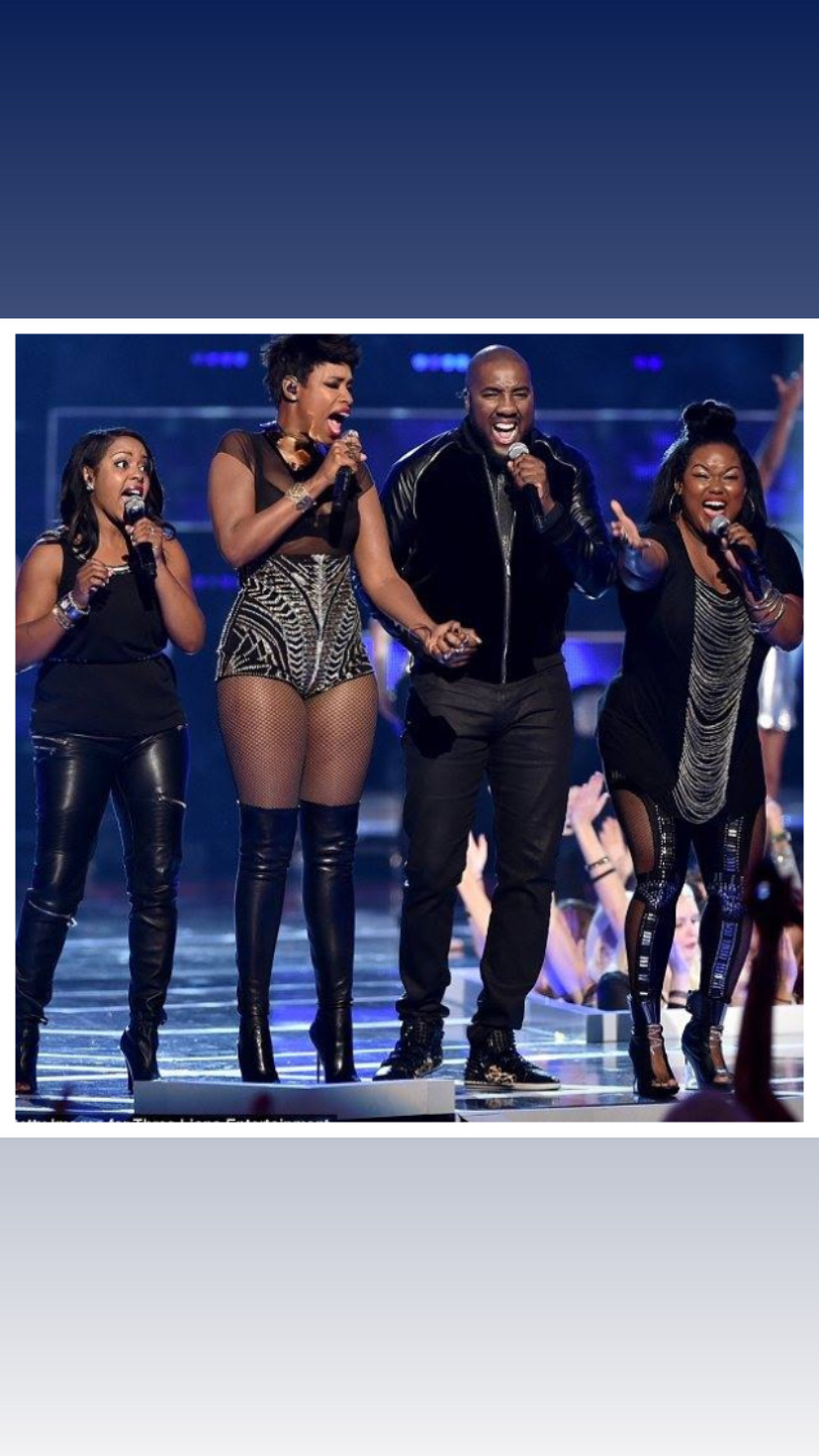 Four performers on stage, three women and one man, singing and dancing with microphones, wearing black outfits, stage lights in background.