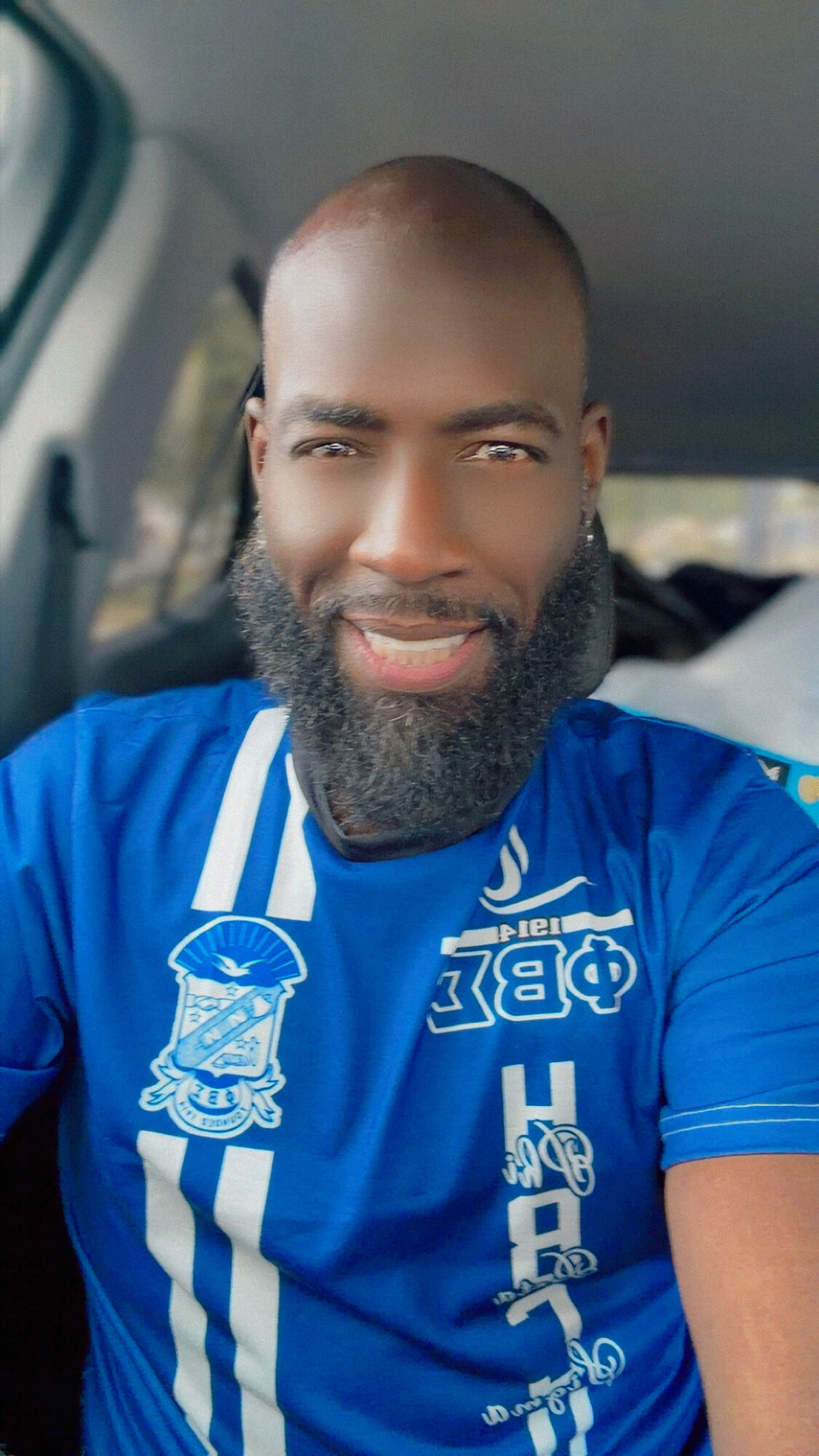 Man with a beard smiling, wearing a blue sports jersey inside a vehicle.