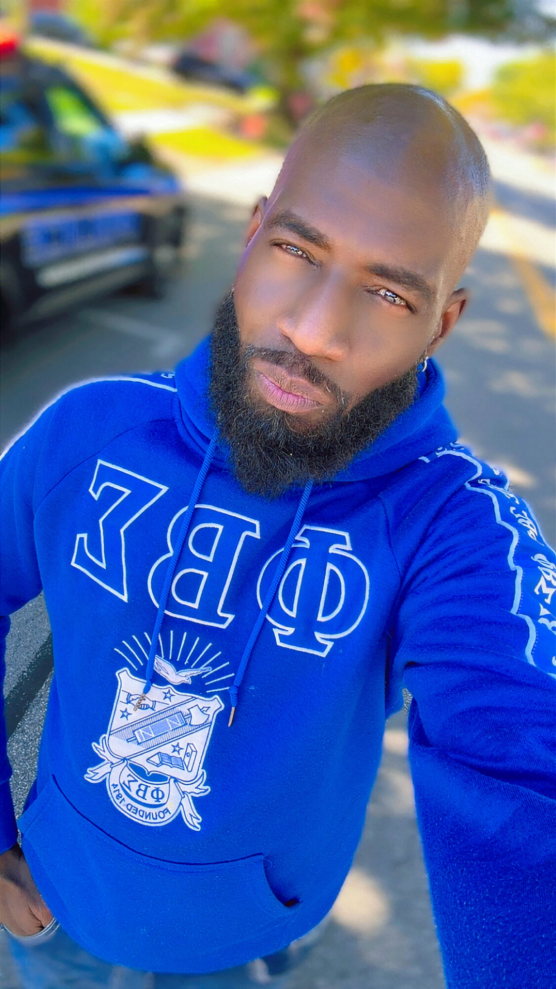 Man with a beard taking a selfie outdoors, wearing a blue hoodie with Greek letters and a crest, sunny day.