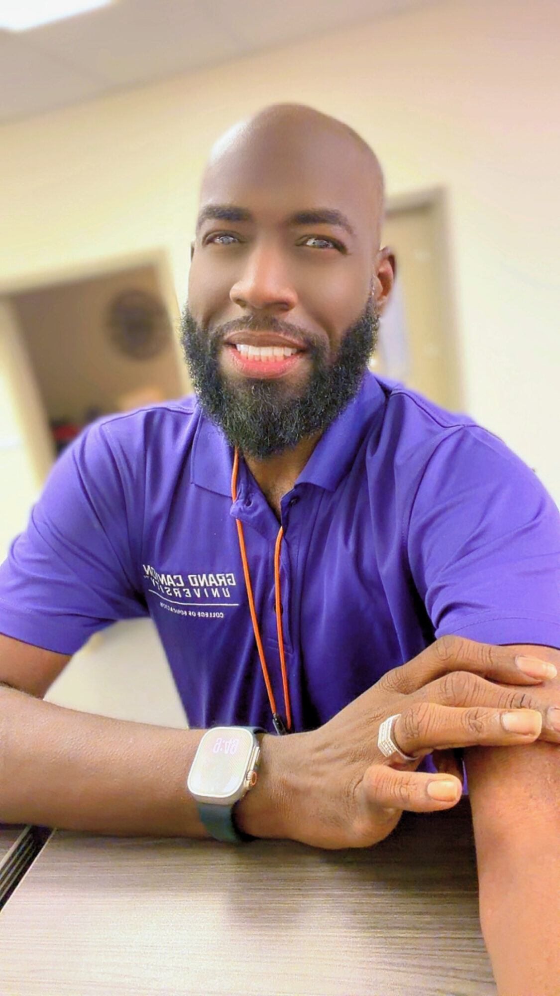 Man with a beard and bald head wearing a purple shirt, sitting at a table, smiling, with a smartwatch on his wrist.