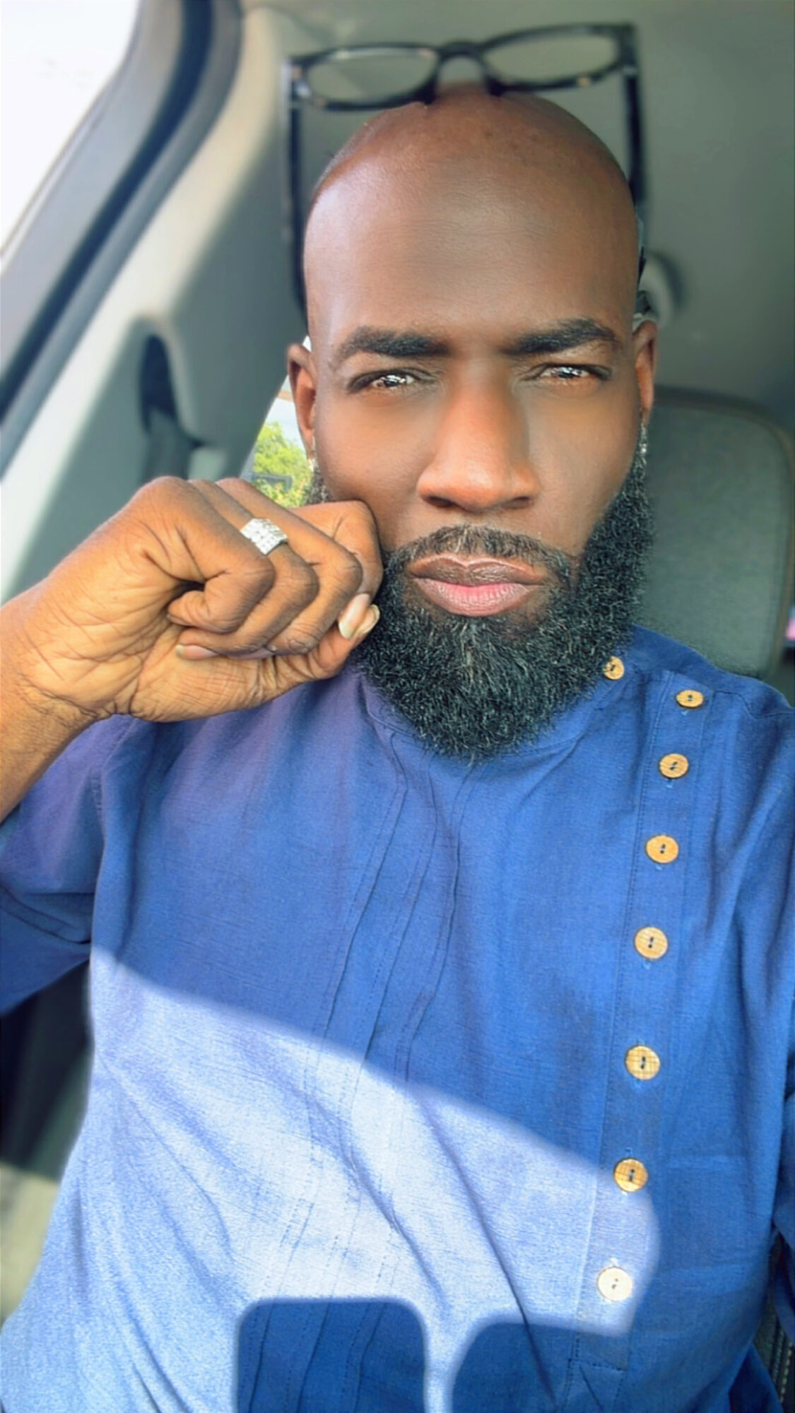 Man with glasses on head, beard, wearing blue shirt, sitting in vehicle, looking at camera, hand near face.