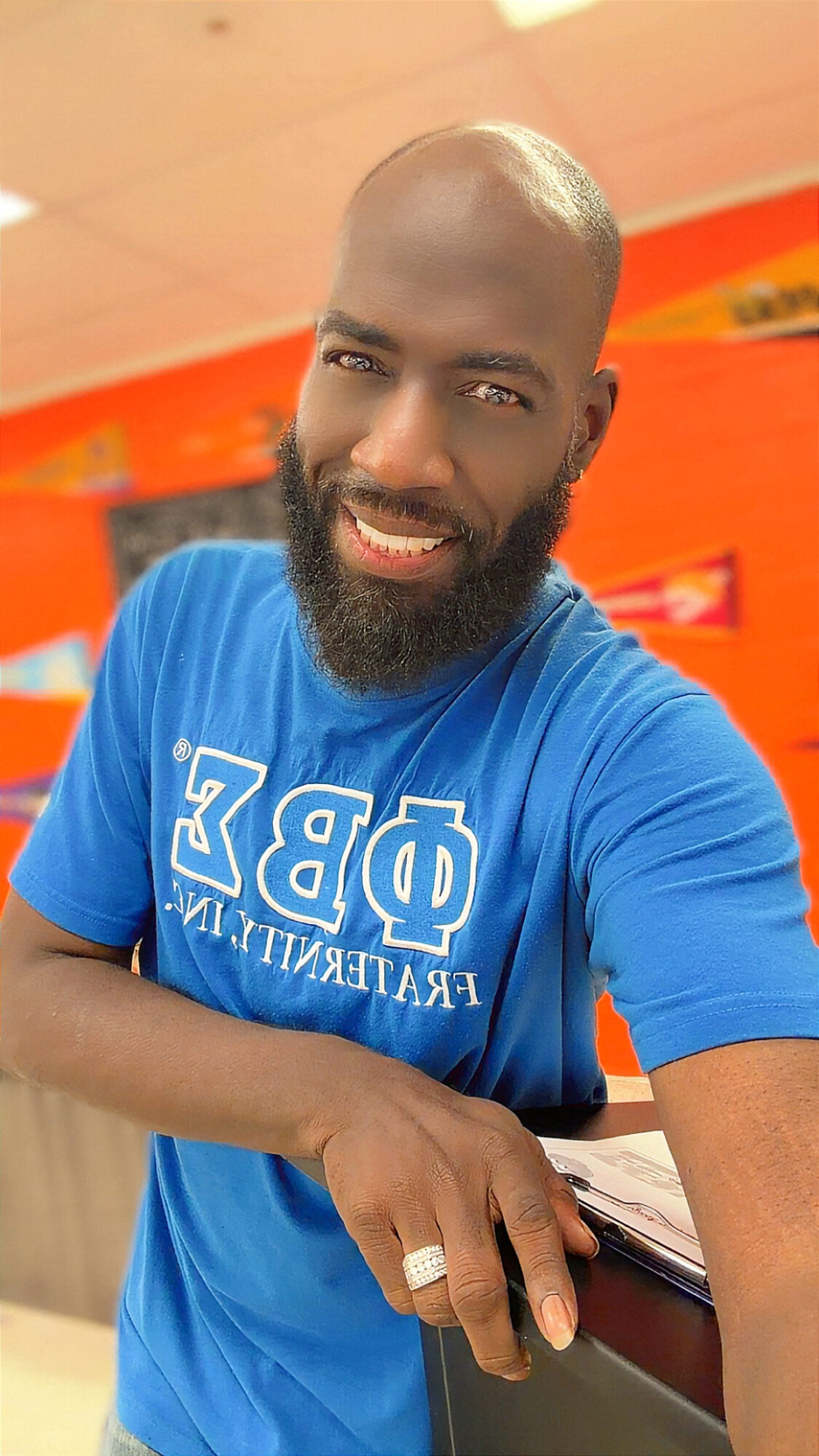 Man with a beard smiling, wearing a blue T-shirt, in an indoor setting with orange and yellow background.