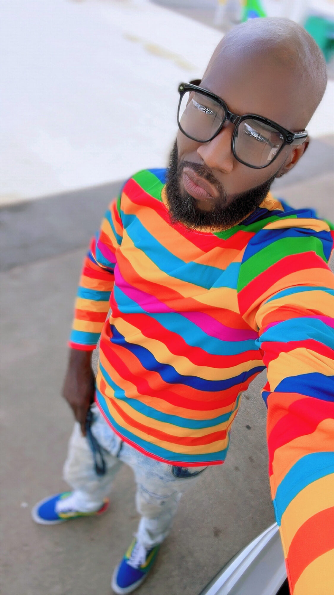Person with glasses and a colorful striped shirt taking a selfie outdoors, with a bright background.