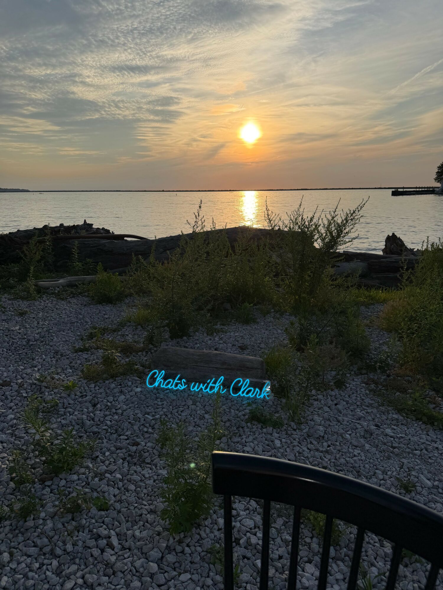 Sunset over a body of water with a cloudy sky, trees, and a gravel path, with a blue sign reading 'Blue and Gold Club'.