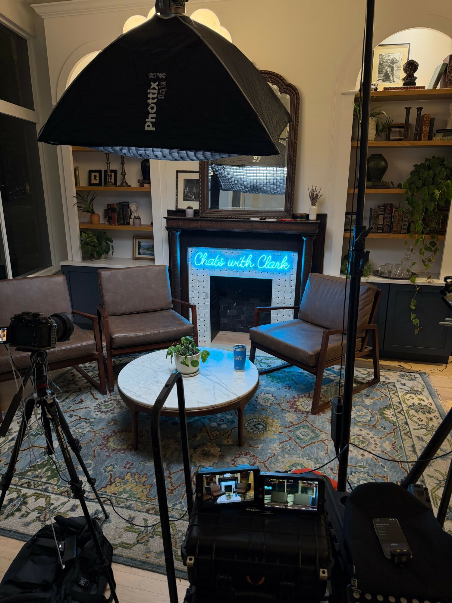 Studio setup with a computer monitor, microphone, and lighting equipment in a cozy room with bookshelves and a patterned rug.