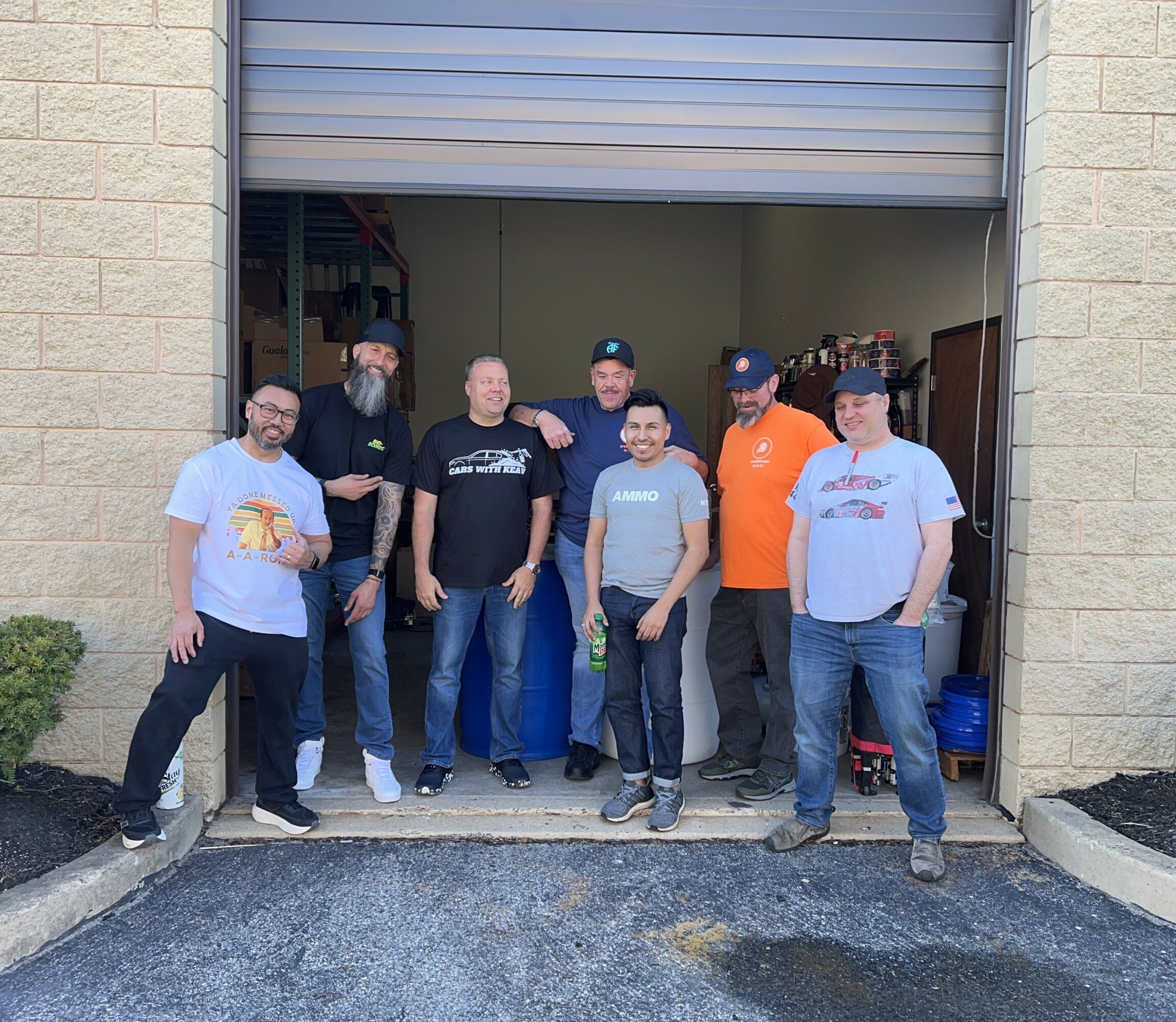 Group of seven people standing in front of an open garage door, some wearing casual clothes and hats.