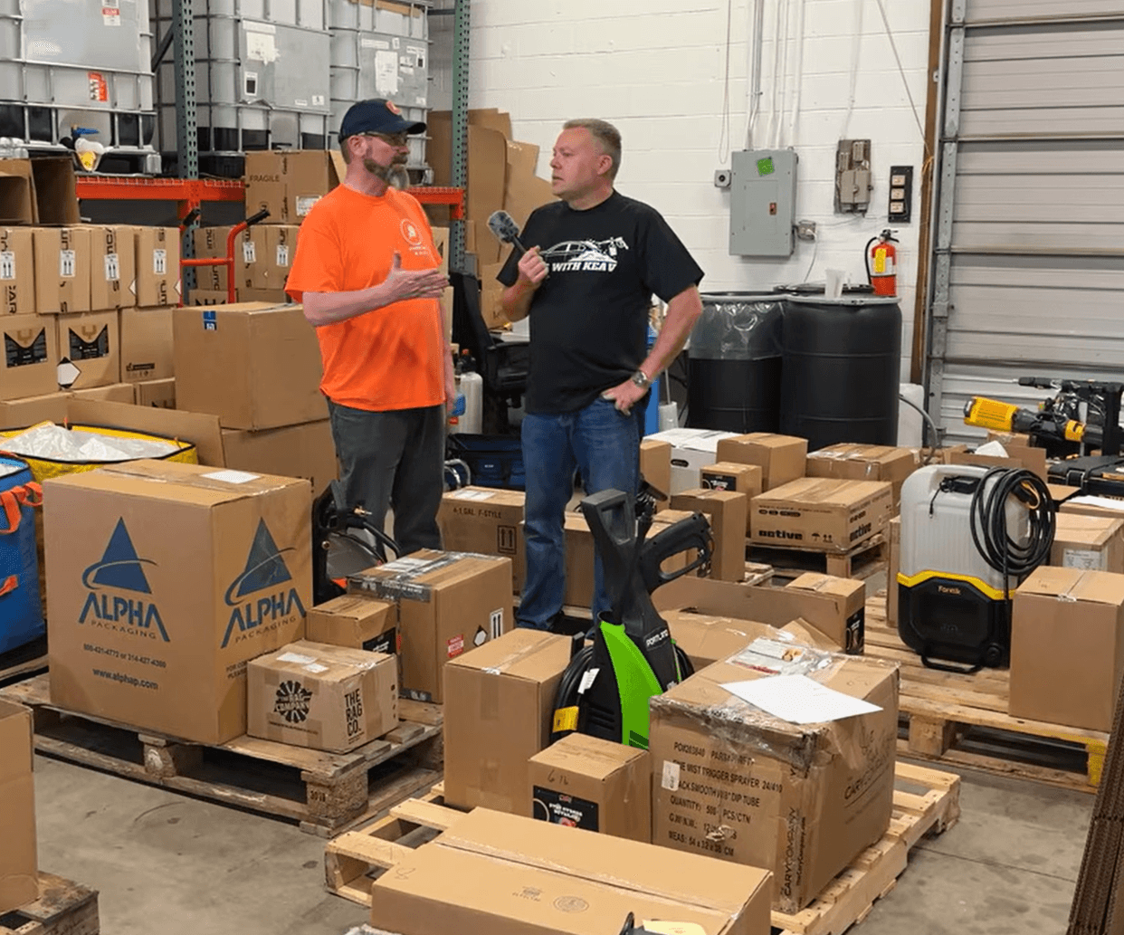 Two men are talking in a warehouse surrounded by boxes and equipment, with shelves and a garage door in the background.