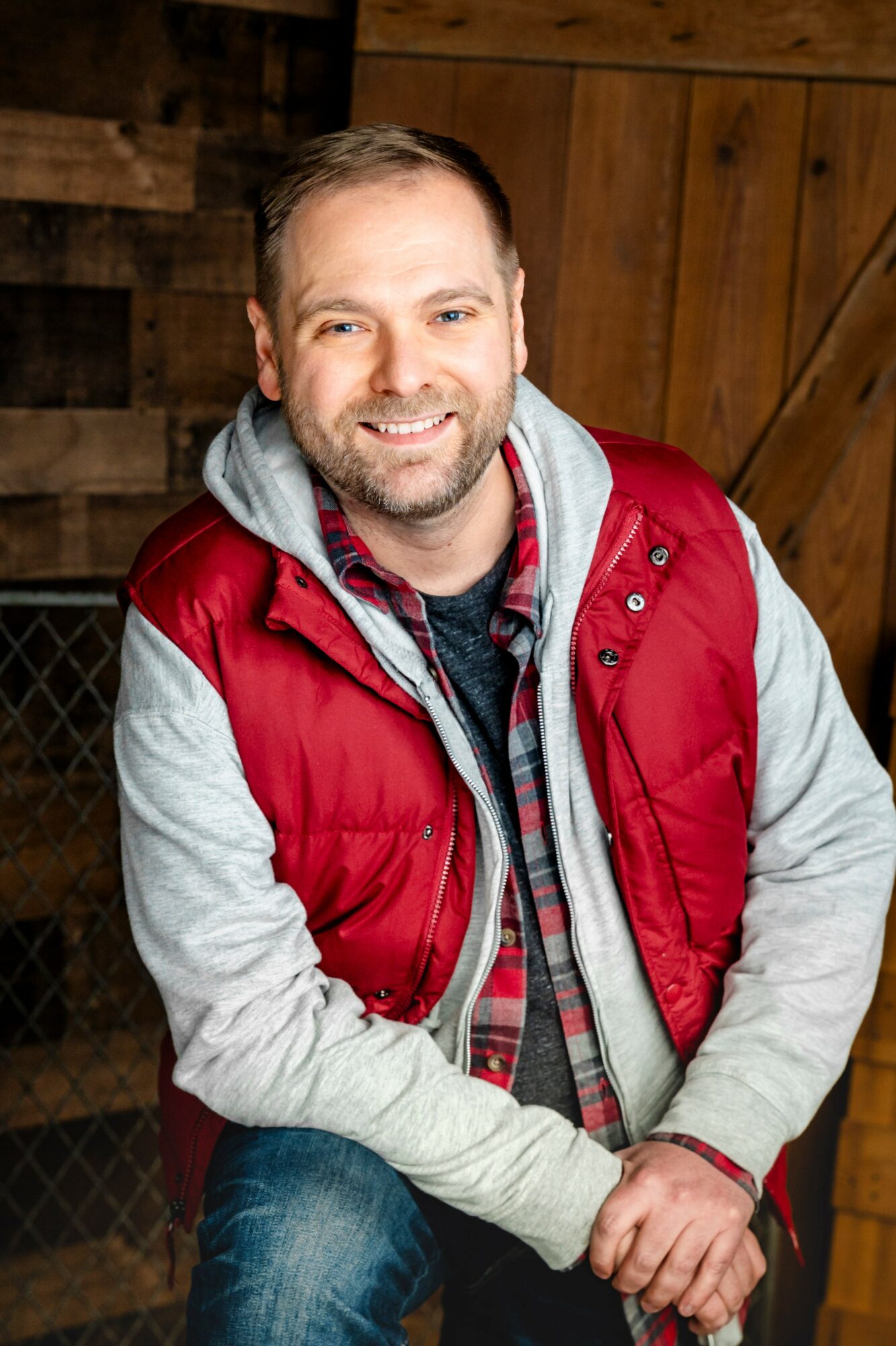 Smiling man with beard wearing a red vest, gray hoodie, and plaid shirt, sitting indoors with wooden background.