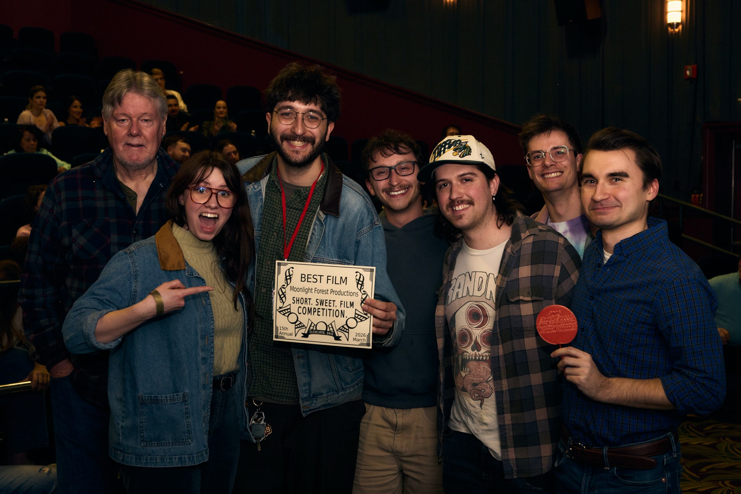 Group of seven people smiling, one holding a certificate, in a theater setting with audience in background.