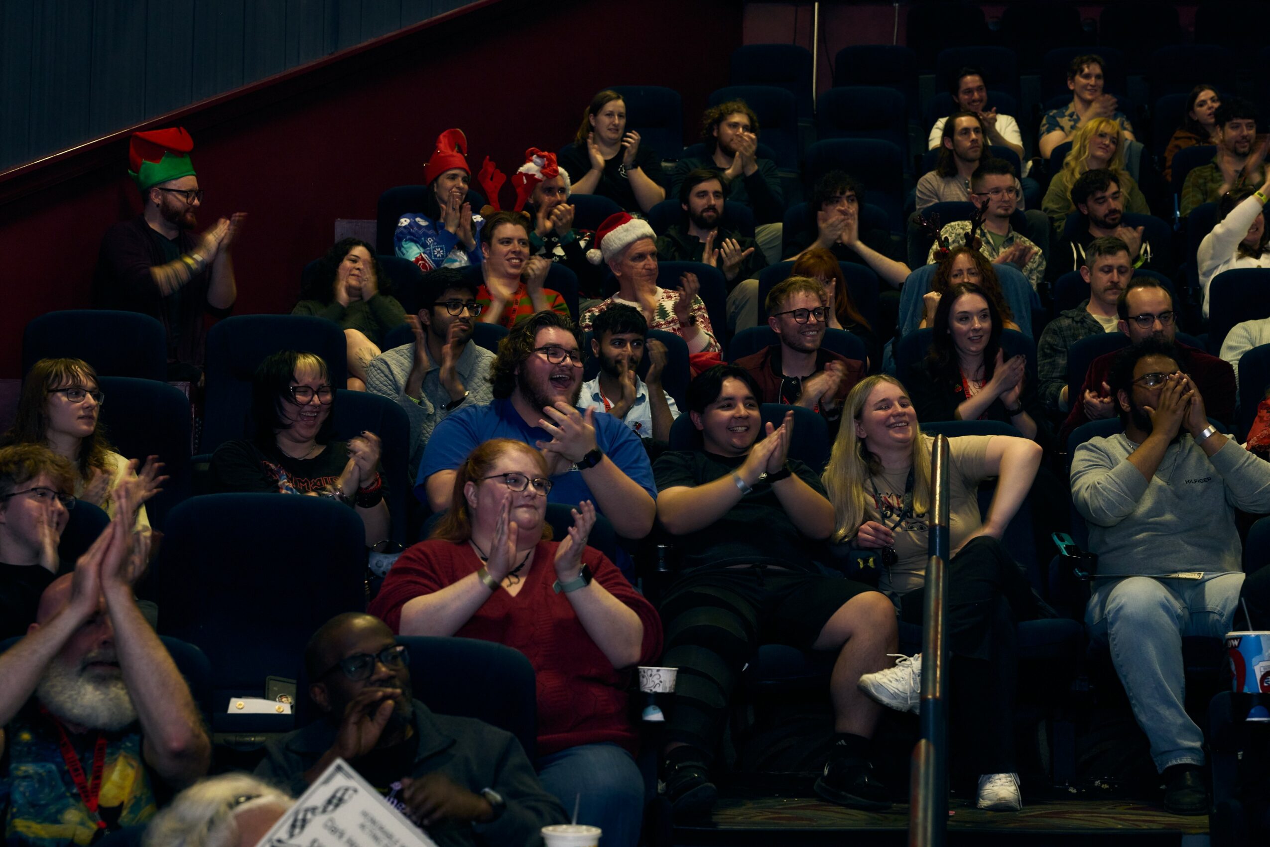 Audience members seated in a dark theater, clapping and smiling, some wearing festive hats and glasses.
