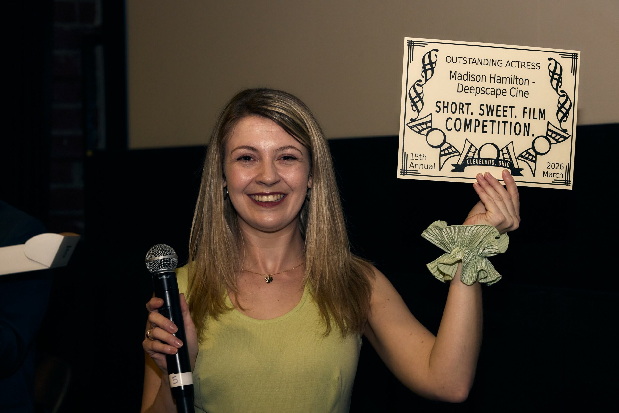 Woman smiling holding a certificate and microphone, standing indoors with a dark background.