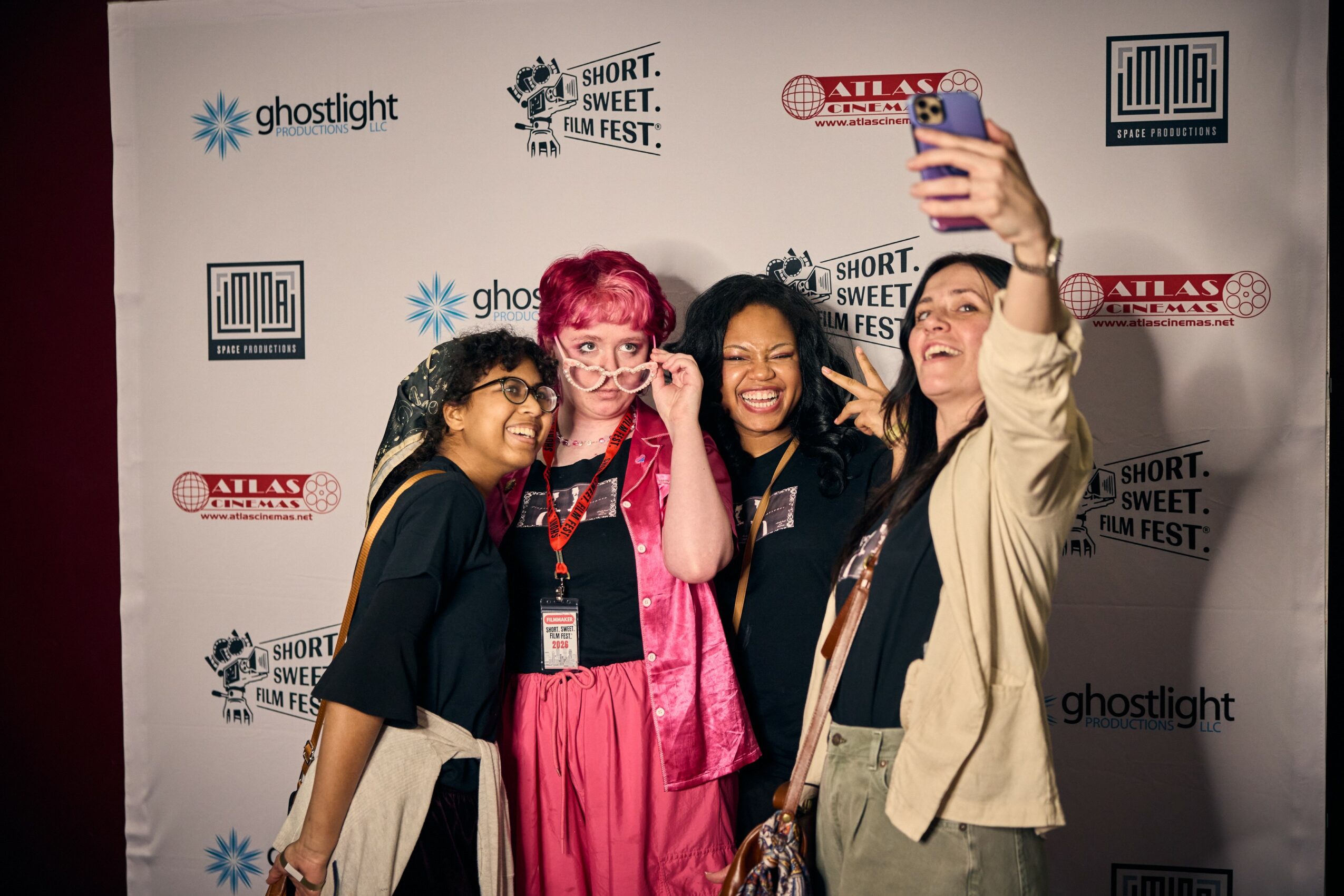 Four women taking a selfie together at an event, smiling and posing in front of a backdrop with logos.