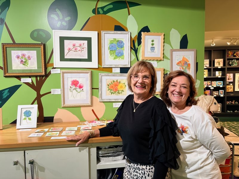 Two women smiling and standing behind a table with framed artwork in a colorful gallery setting.