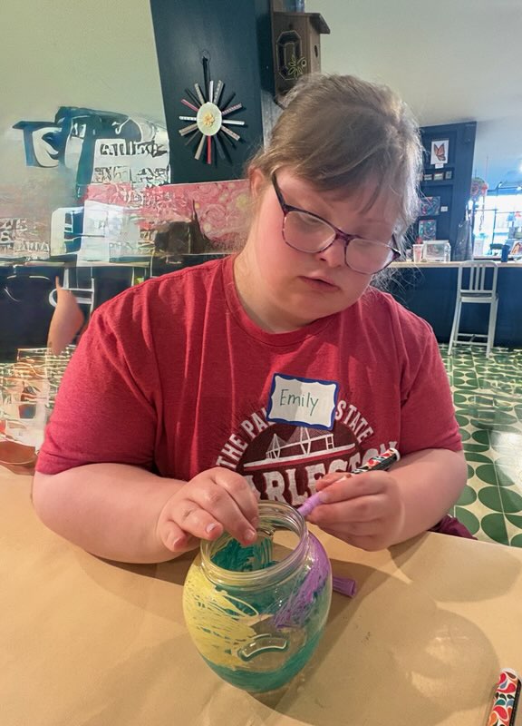 Girl with glasses and a red shirt looks at a paper in a decorated jar on a table.