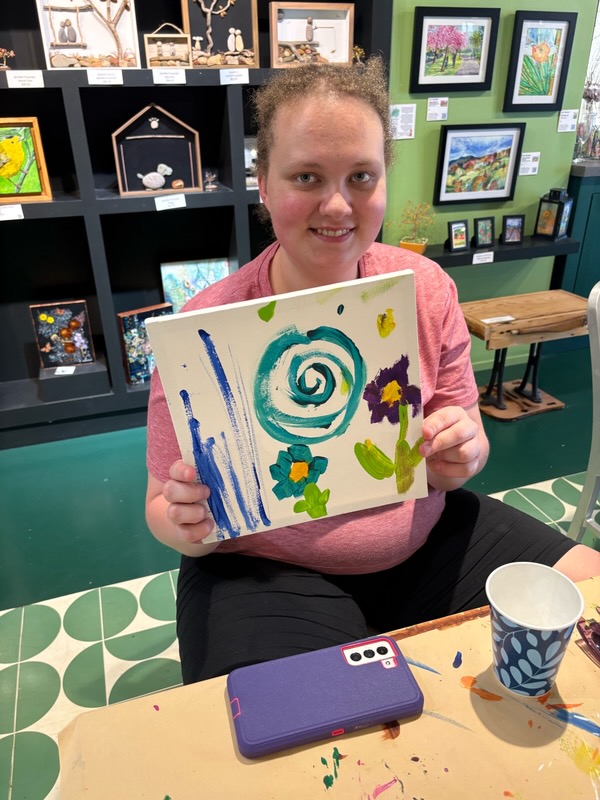 Young girl holding a colorful abstract painting with swirls and shapes, sitting at a table in an art gallery or classroom.