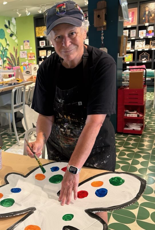 Person wearing a cap and apron, decorating a large butterfly-shaped cake with colorful dots, in a decorated room.