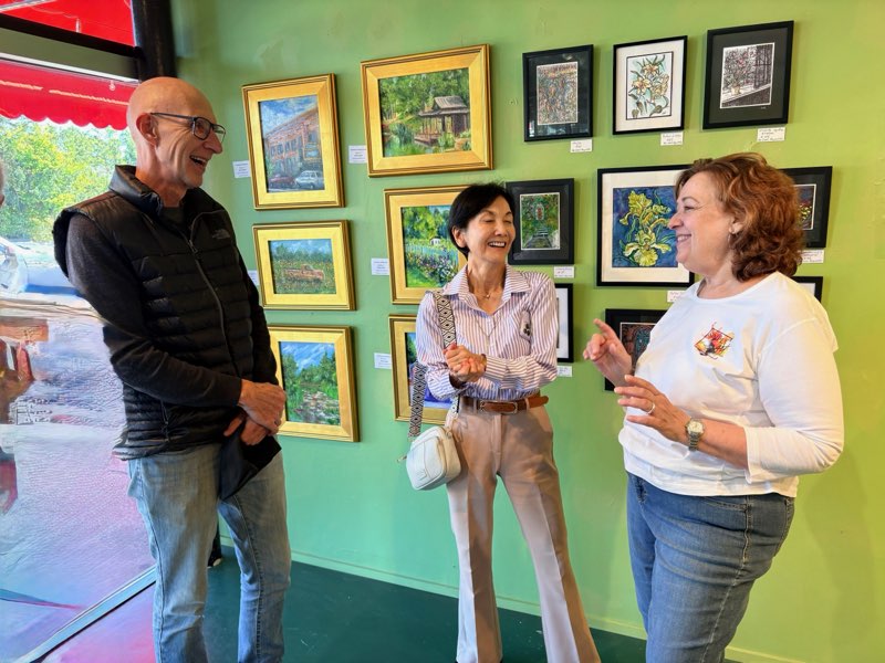 Three people smiling and talking in an art gallery with framed paintings on a green wall.