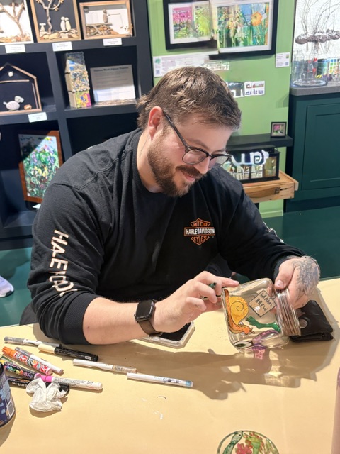 Man with glasses and beard, wearing a black shirt, sitting at a table, looking at a comic book, in a colorful room.