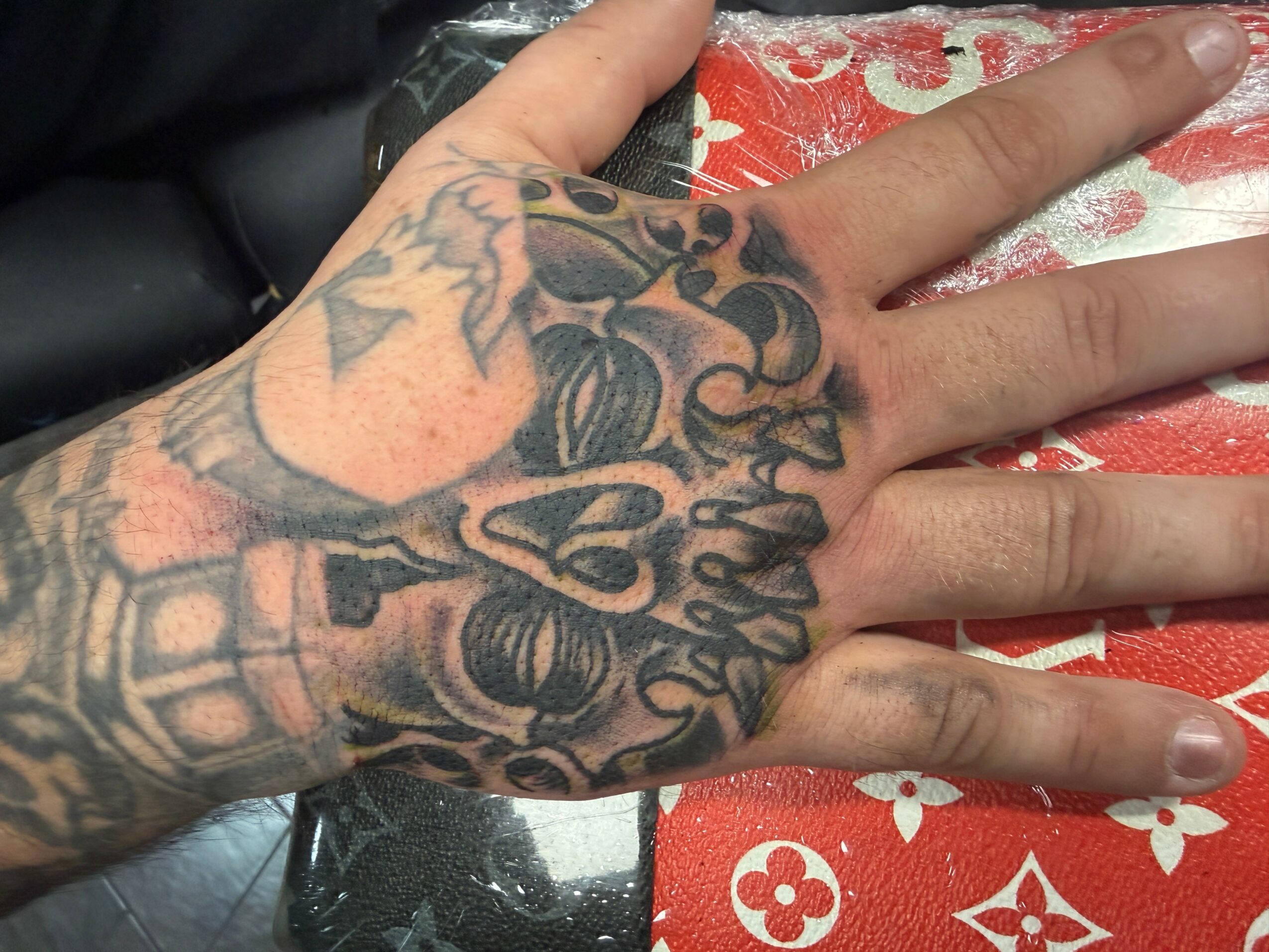 Tattooed hand with a detailed design covering the back of the hand and fingers, resting on a red and white patterned surface.